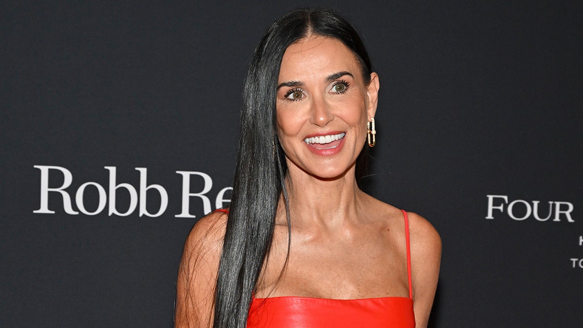 Demi Moore in a red latex strap dress looks to the left at the carpet and smiles