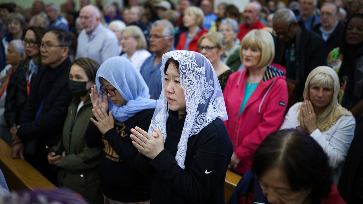 An Asian woman wearing a veil praying next to a bunch of other people who are also praying.