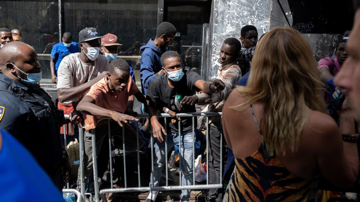 Migrants outside NYC hotel on sidewalk behind barrier