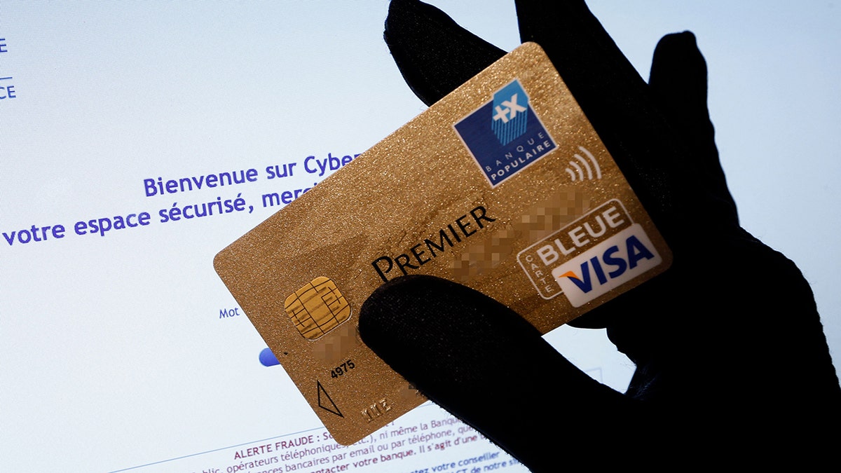 Uma imagem mostra uma pessoa segurando um cartão de crédito Visa Premier em frente a uma tela de computador exibindo um site da Internet com uma mensagem de alerta contra fraudes. 