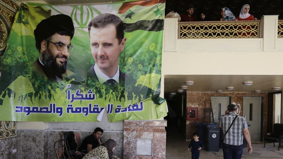 Syrer versammeln sich in der Lobby eines beschädigten Wohnblocks und tragen ein Plakat mit dem syrischen Präsidenten Baschar al-Assad (rechts) und Hassan Nasrallah, dem Chef der schiitischen Bewegung Hisbollah im Libanon, nach einer Autobombe in der Nähe des verehrten schiitischen Schreins Sayyida Zeinab (im Süden). der syrischen Hauptstadt Damaskus am 25. April 2016.