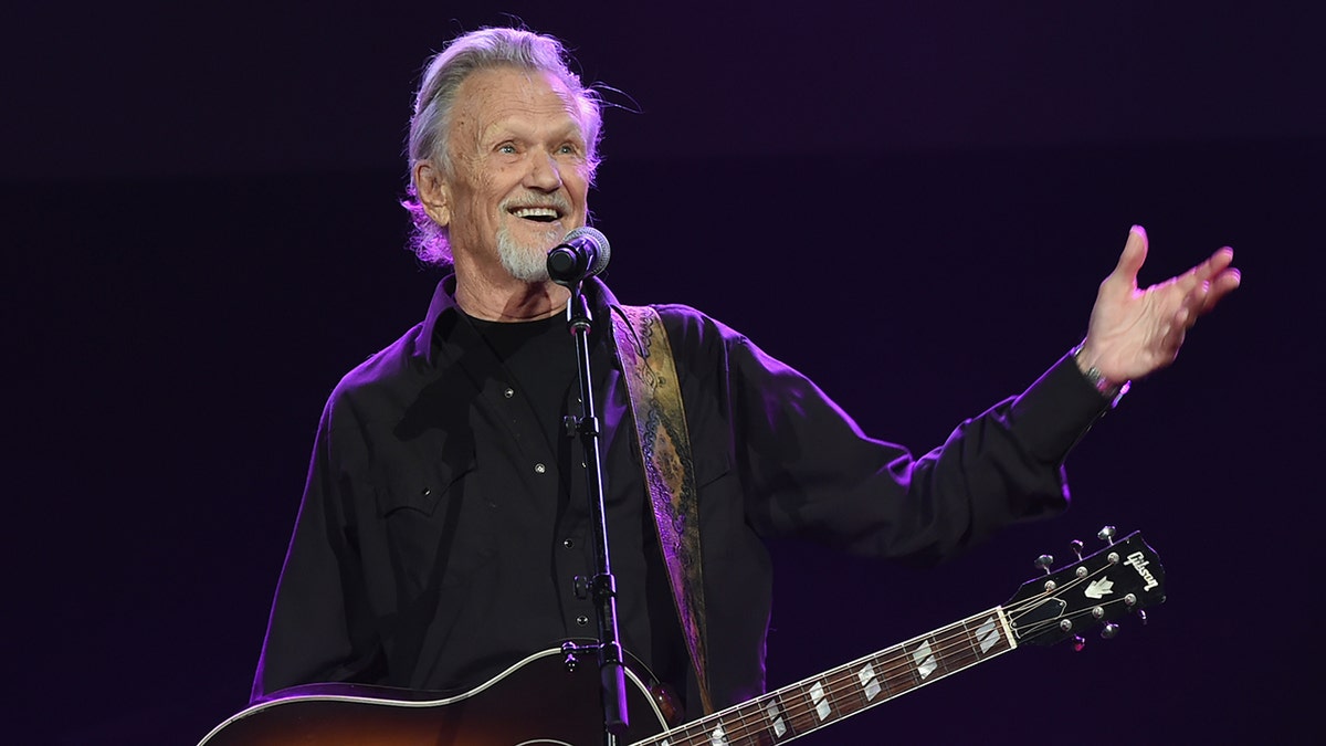 Kris Kirstofferson tersenyum dalam kemeja hitam di atas panggung dengan gitar di bahunya