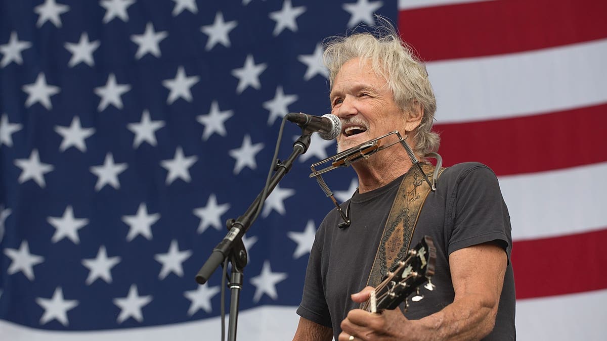 Kris Kristofferson singing and playing guitar in front of American flag