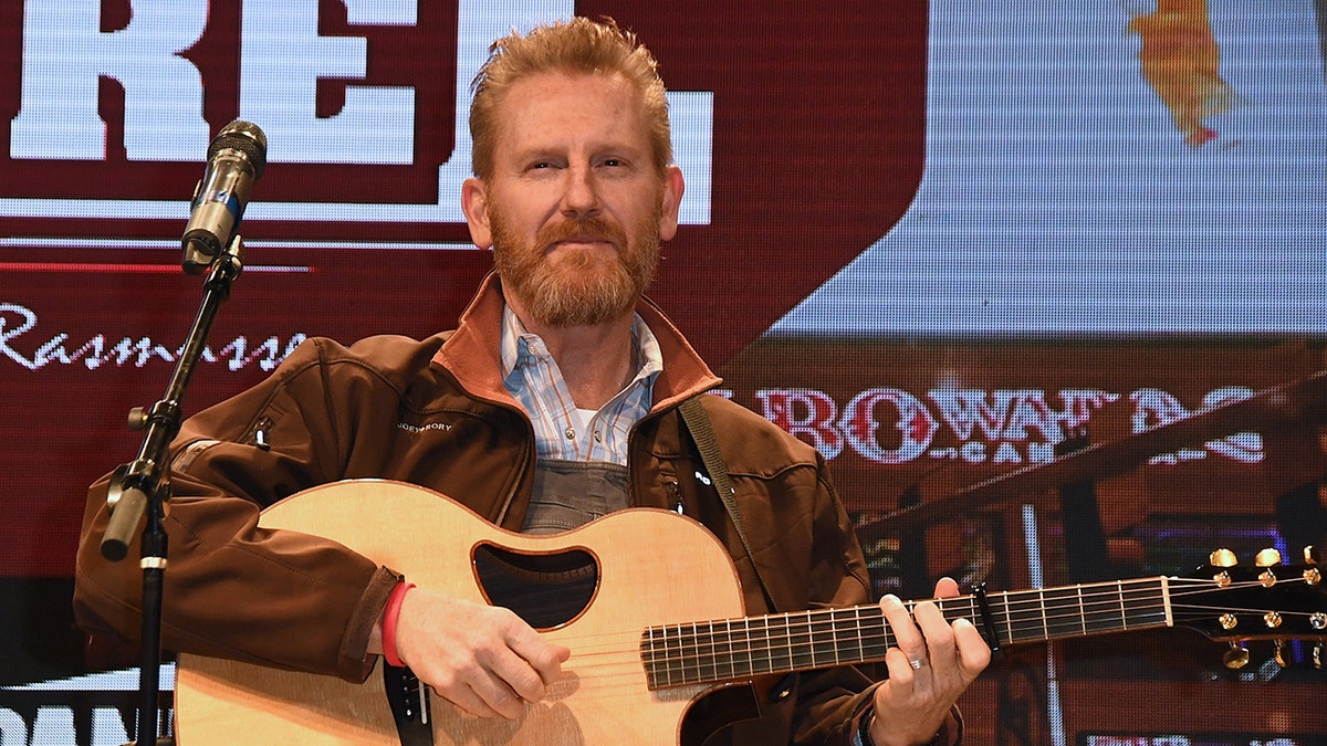 Rory Feek con una chaqueta marrón mira hacia arriba en el escenario mientras toca la guitarra