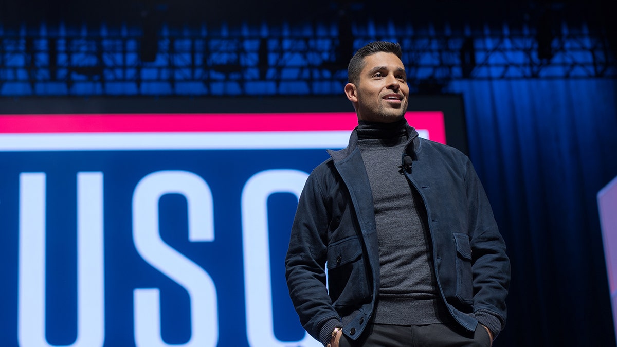 Wilmer Valderrama on stage wearing a grey turtleneck and a navy jacket with a USO sign behind him.