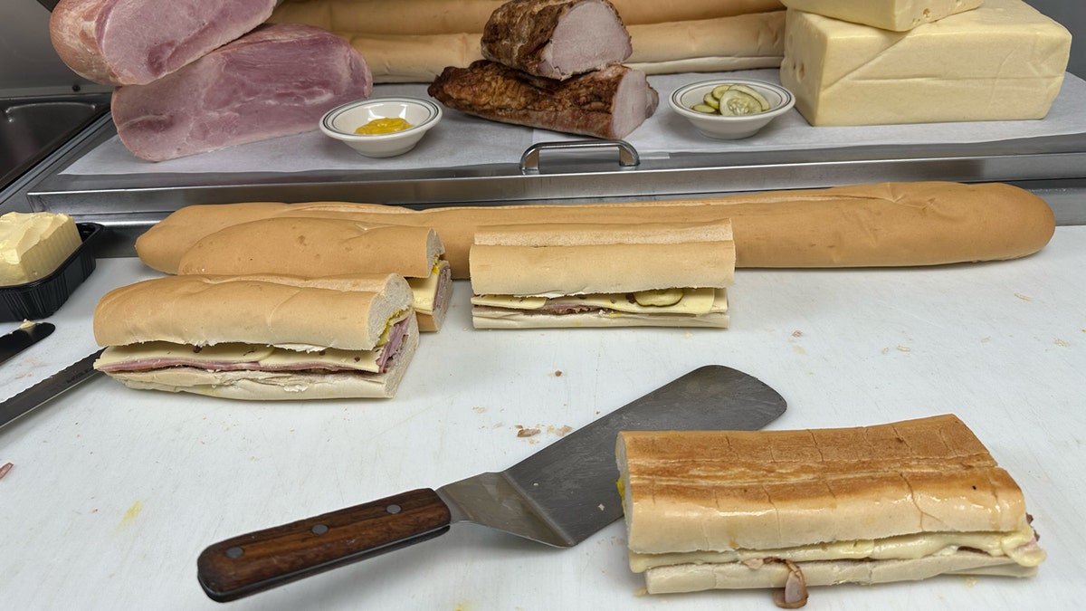 Un sándwich cubano recién prensado de Versalles se sienta en una tabla de cortar junto a otros sándwiches sin prensar en la cocina del restaurante de Miami, 26 de septiembre de 2024.