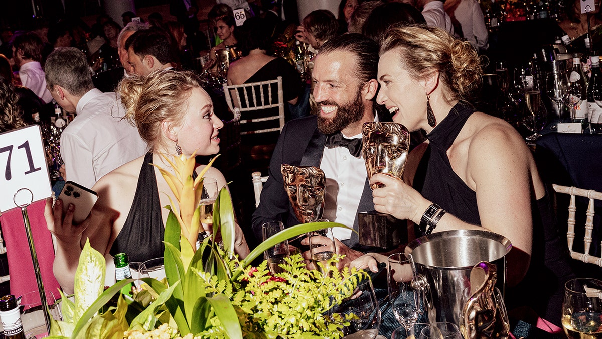 Mia Threapleton, con un vestido negro, sentada frente a su madre Kate Winslet, con un vestido halter negro, y su marido Edward Abel Smith, con un esmoquin clásico, en los BAFTA