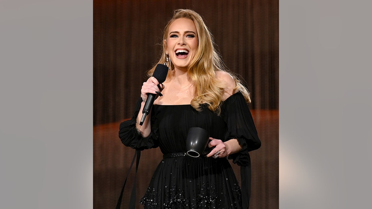 Adele in a black off the shoulder dress laughs on stage holding the microphone