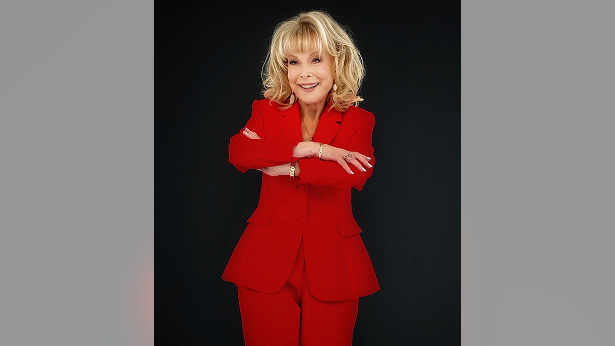 Barbara Eden dengan setelan celana merah menyilangkan lengannya dan tersenyum