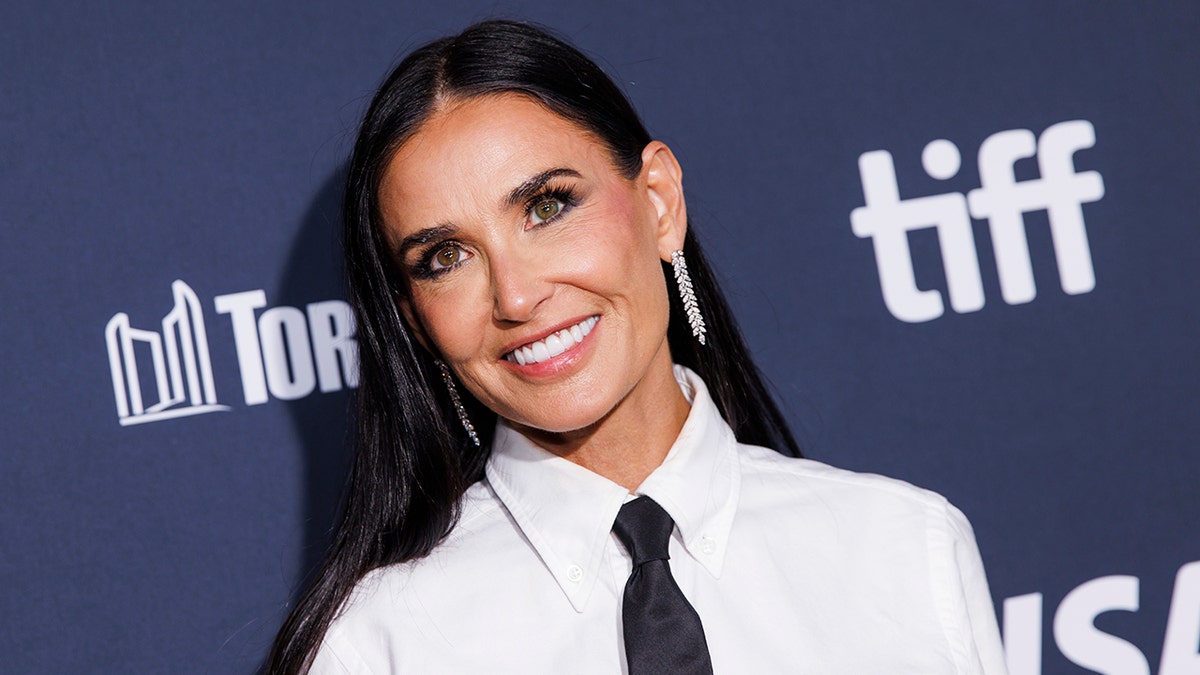 Demi Moore in a white blouse with a black tie tilts her head and smiles