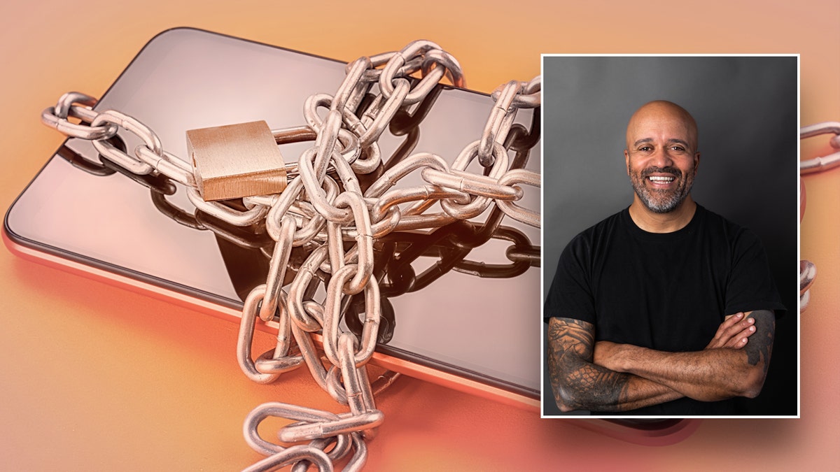 A photo of Carlos Whittaker, in which he is a bald man with a beard and wearing a black shirt, holding a phone wrapped around a chain.