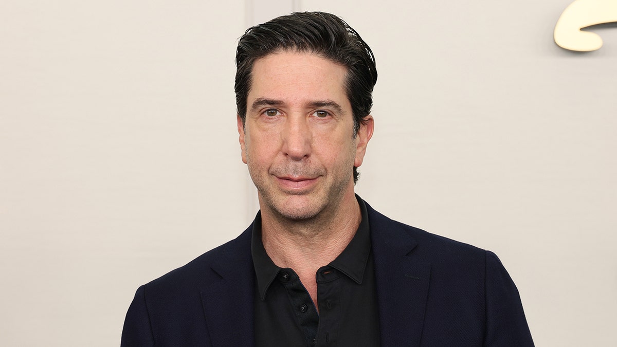 David Schwimmer con traje y camisa negra se muestra serio en la alfombra