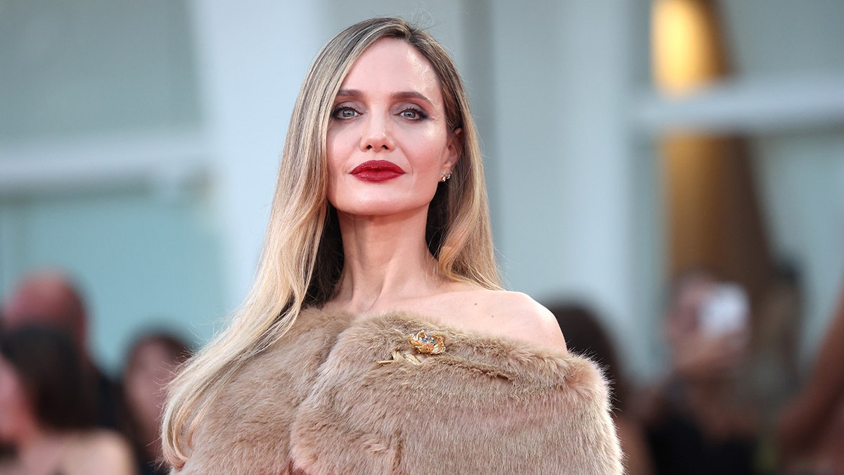 Angelina Jolie with a red lip has an off-the-shoulder fur jacket on in Venice