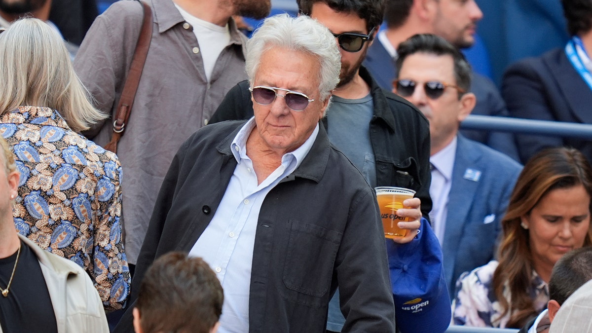 Dustin Hoffman berjalan ke tempat duduknya di AS Terbuka dengan mantel abu-abu dan kemeja berkancing biru