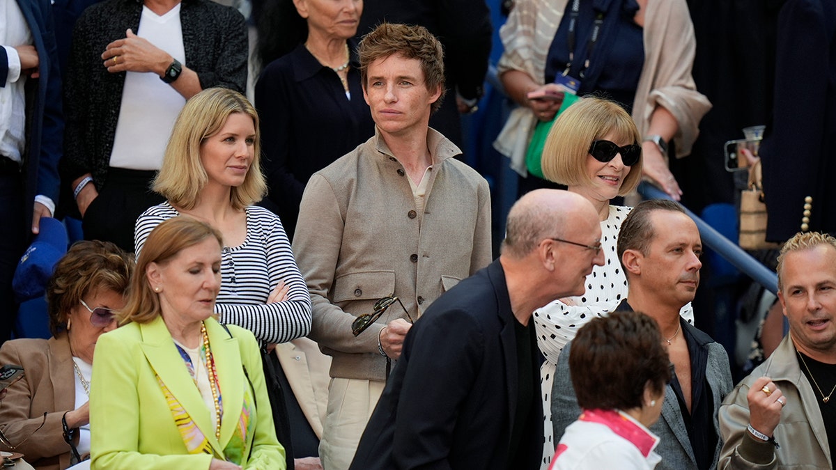 Eddie Redmayne dengan kemeja cokelat duduk di sebelah Anna Wintour di AS Terbuka dan Billie Jean King duduk di bawah mereka dengan punggungnya menghadap kamera