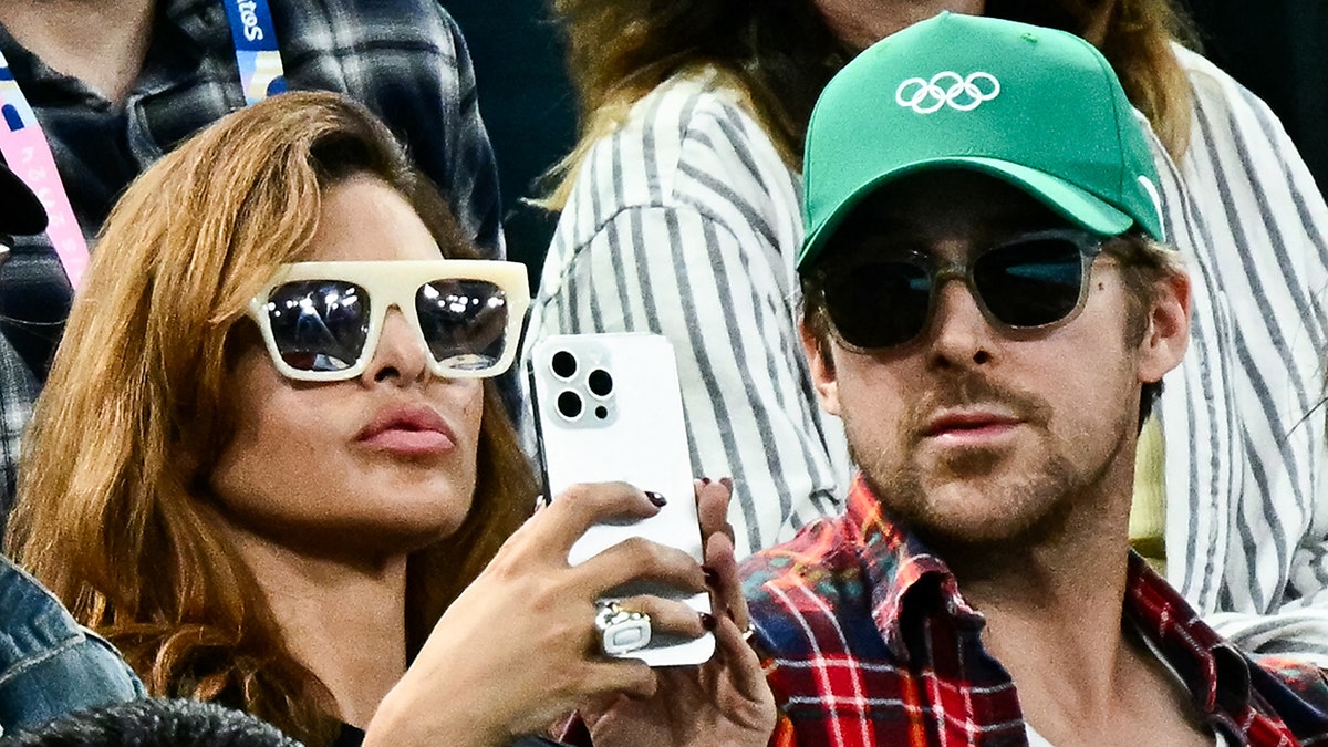 Eva Mendes segura um iPhone branco enquanto usa óculos escuros creme, sentada ao lado do marido Ryan Gosling, com uma camisa xadrez e chapéu verde no US Open