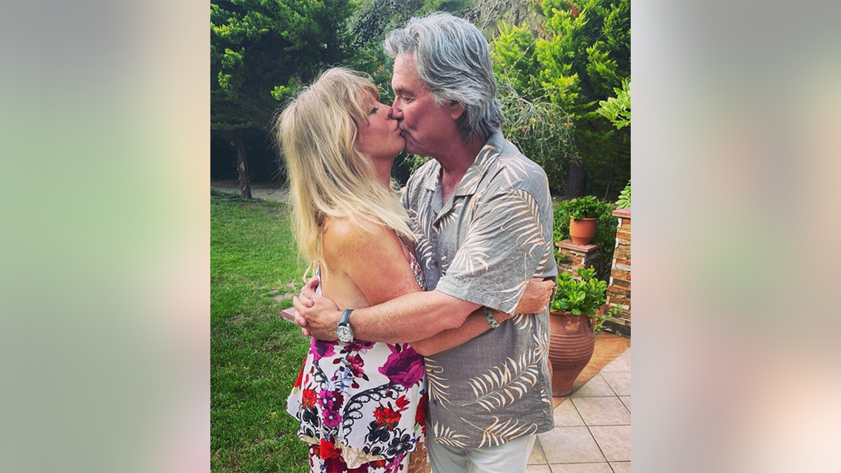 Goldie Hawn con un vestido estampado abraza y besa a Kurt Russell con una camisa gris estampada