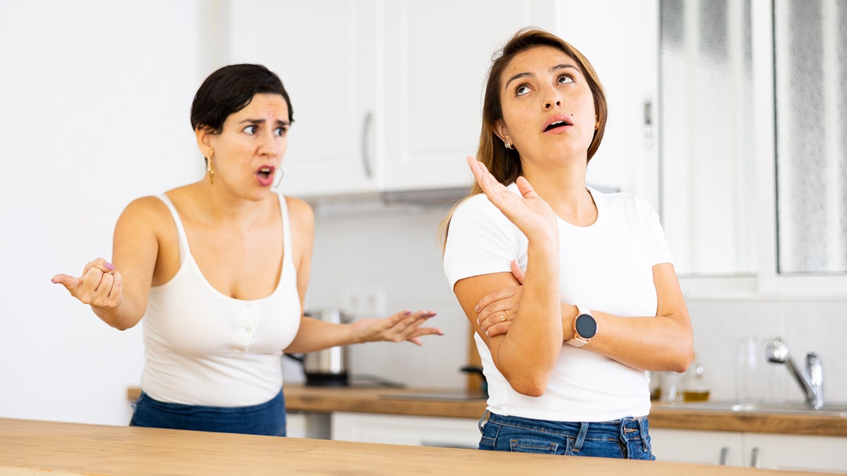 A young Hispanic woman frowns as she stands in her kitchen at home, listlessly listening to the reprimands of her dissatisfied sister