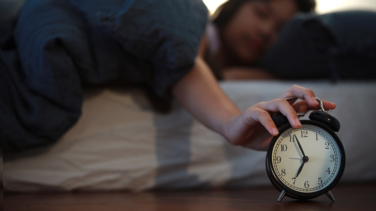 A young Asian woman lies in bed trying to turn off the alarm clock in the bedroom of her home.