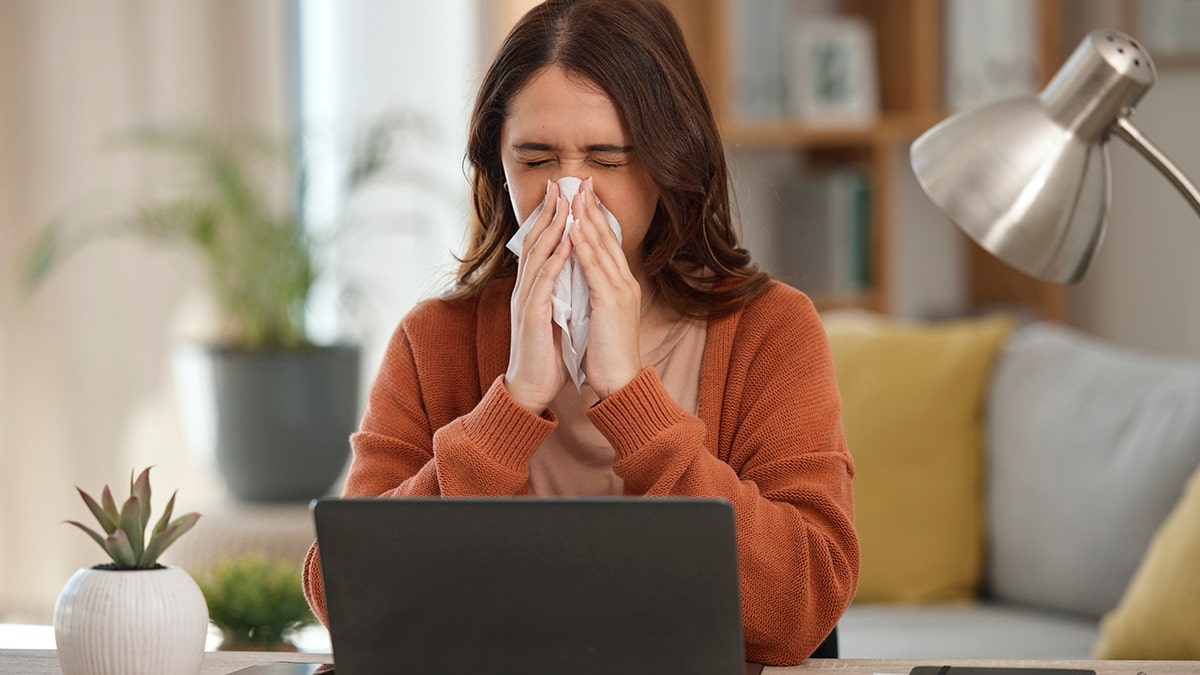 Woman blowing her nose in front of her laptop