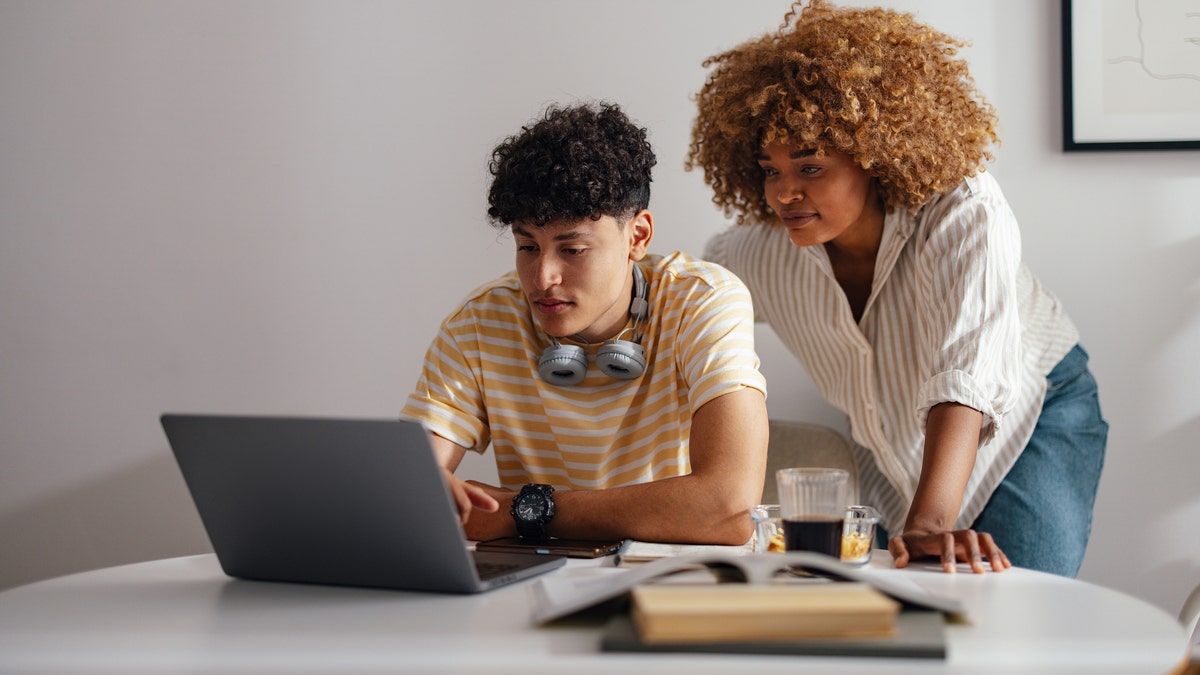 A scene of a mother and her teenage son focusing on a laptop, working together in a well-lit, modern home setting.