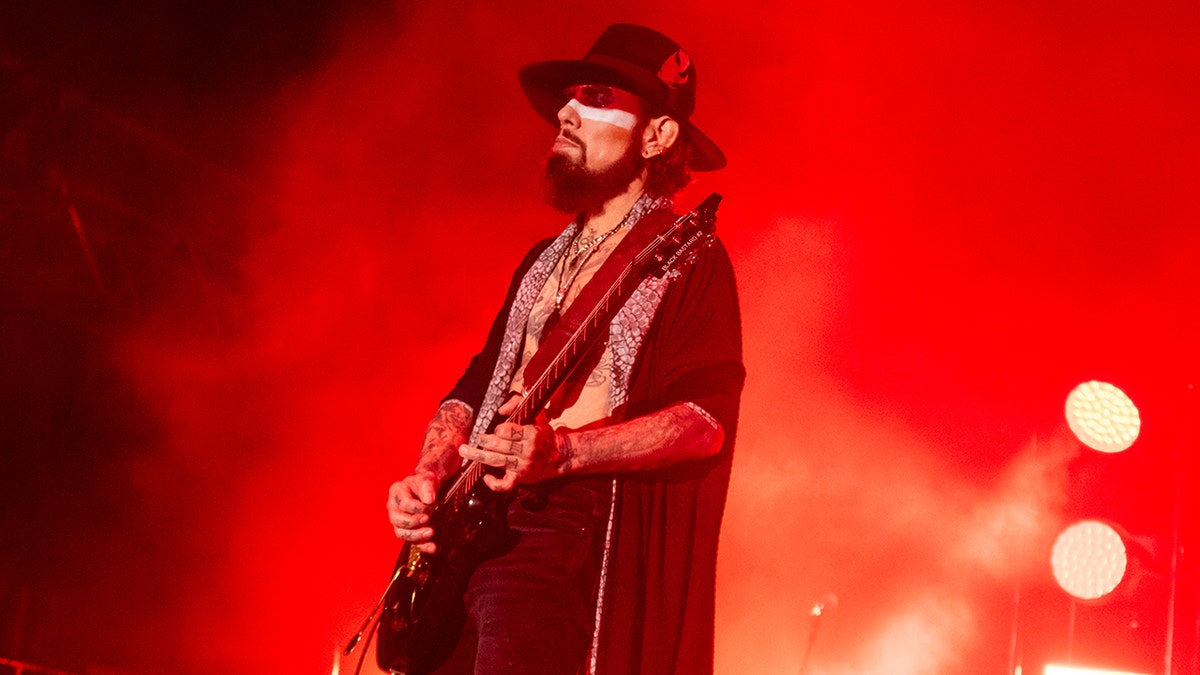 Dave Navarro de camisa preta toca violão no palco com um tom vermelho na foto