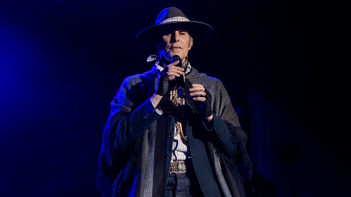 Perry Farrell in a black hat and jacket stands on stage with a microphone.