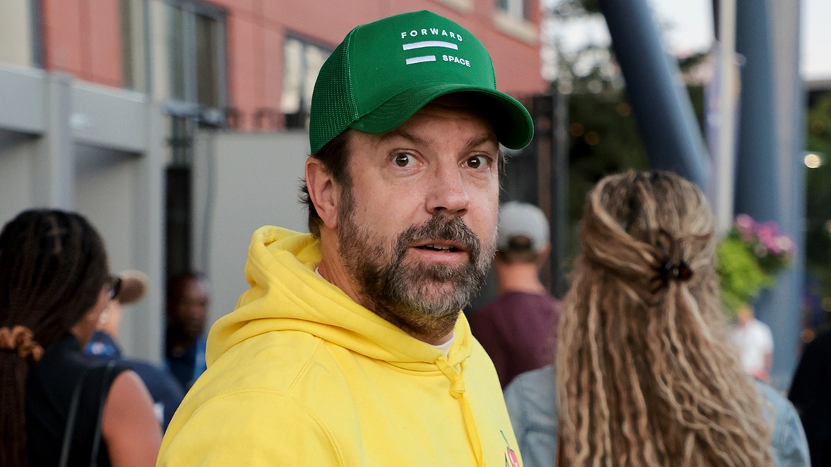 Jason Sudeikis lleva una sudadera amarilla brillante y una gorra verde fuera del US Open, parece un poco asustado por la cámara