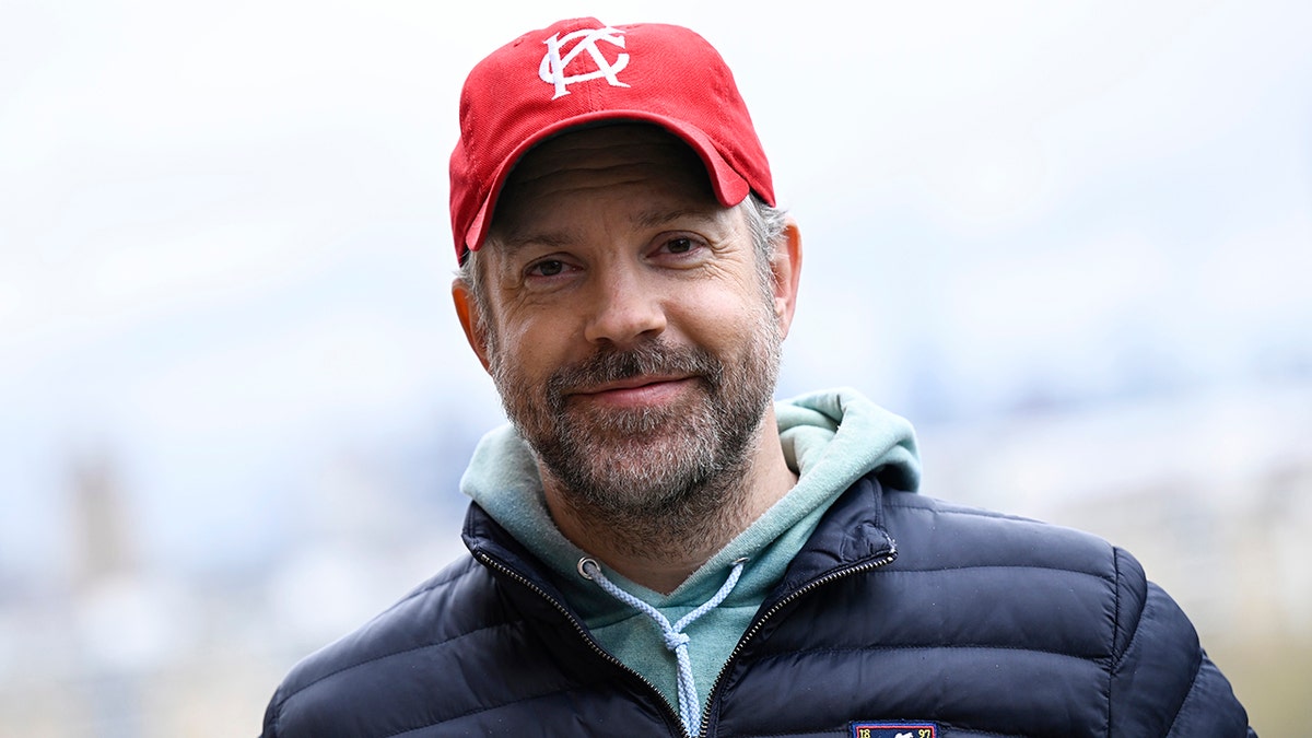 Jason Sudeikis sonríe suavemente llevando una gorra de béisbol roja, una sudadera turquesa y una chaqueta azul abullonada en Londres