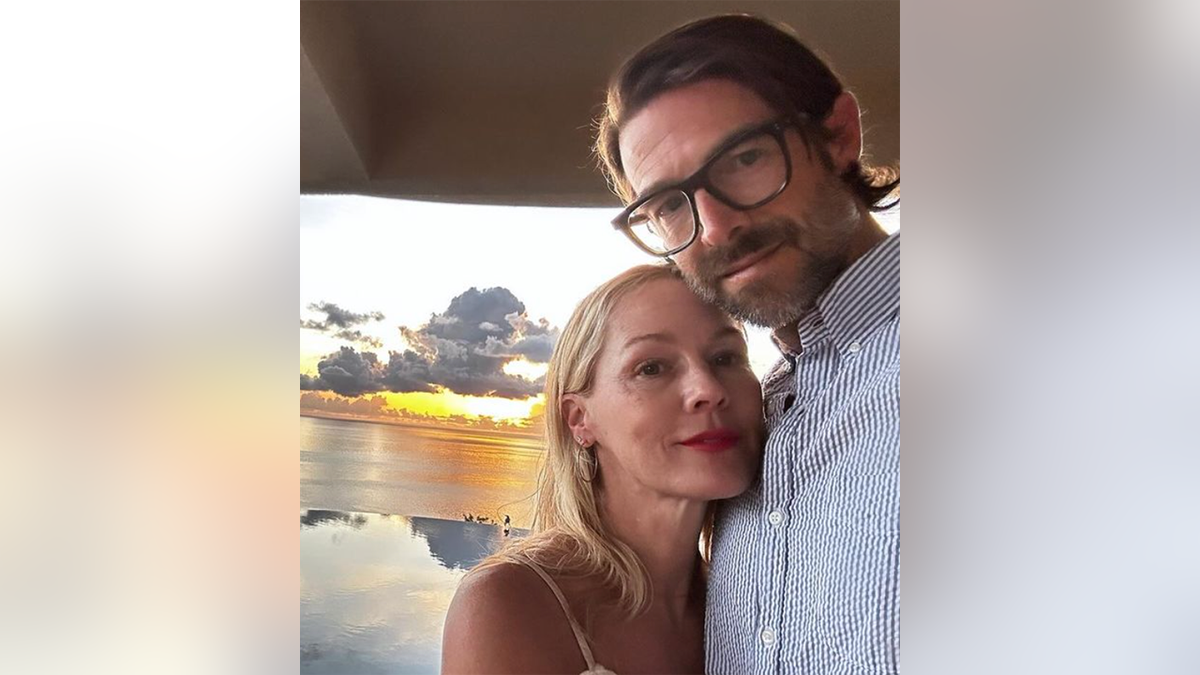 Jennie Garth tersenyum lembut saat dia bersandar pada suaminya Dave Abrams dengan kemeja bermotif dengan matahari terbenam yang menakjubkan di latar belakang