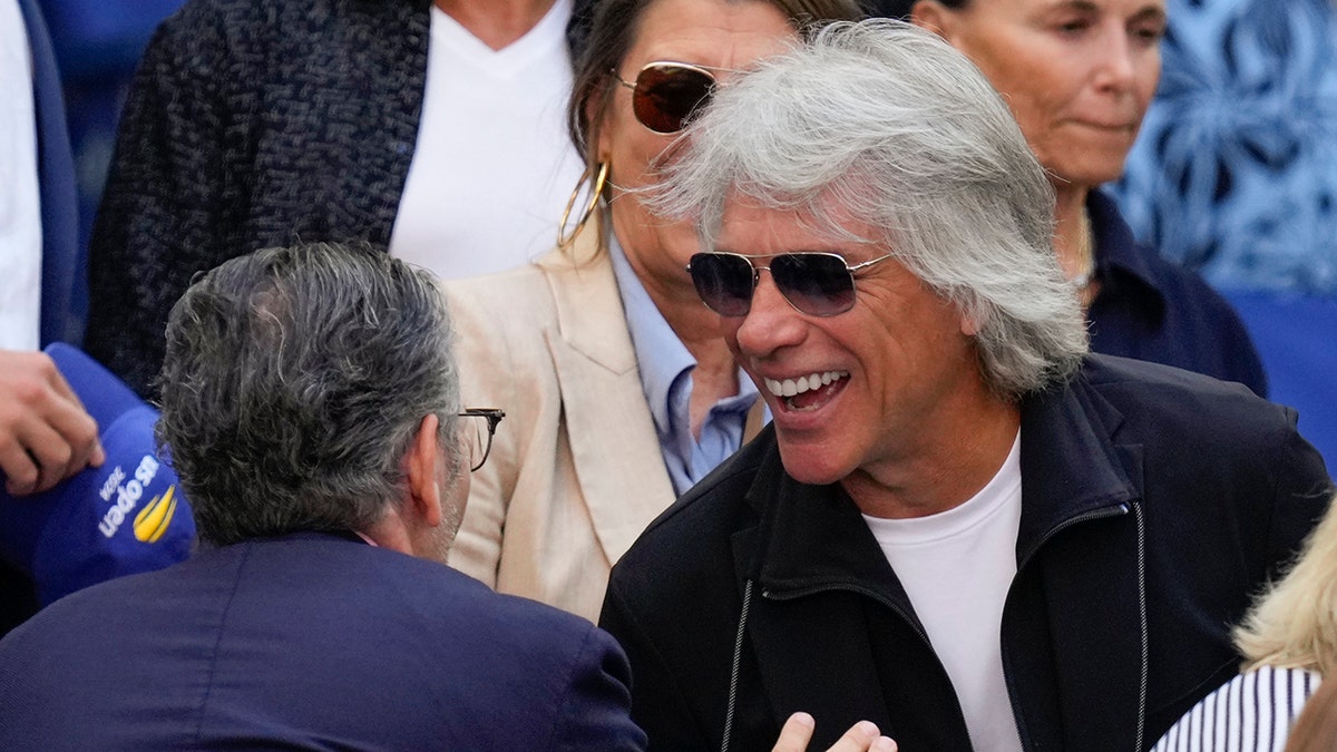 Jon Bon Jovi smiles as he greets someone at the US Open final. He is wearing a black jacket and a white t-shirt