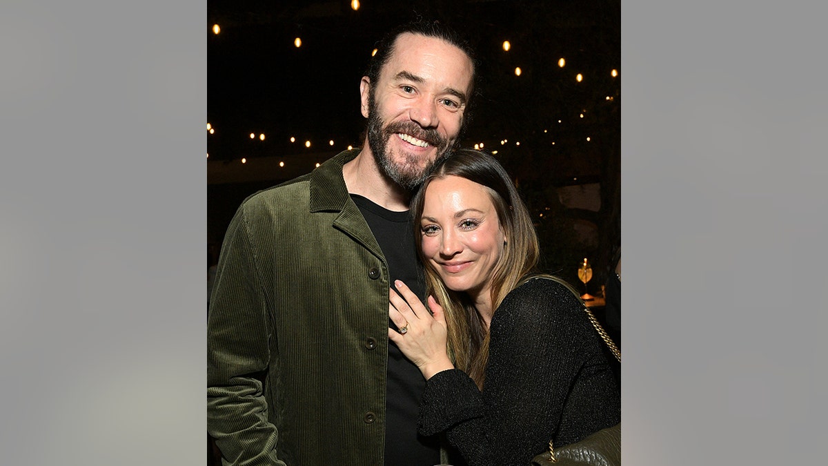 Tom Pelphrey in an olive green jackeet smiles as Kaley Cuoco in black places her hand on his chest