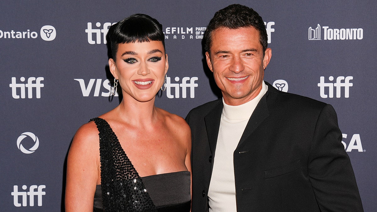 Katy Perry in a one-shoulder sparkly black gown smiles ont the carpet with Orlando Bloom in a white turtleneck and black jacket at the Toronto International Film Festival