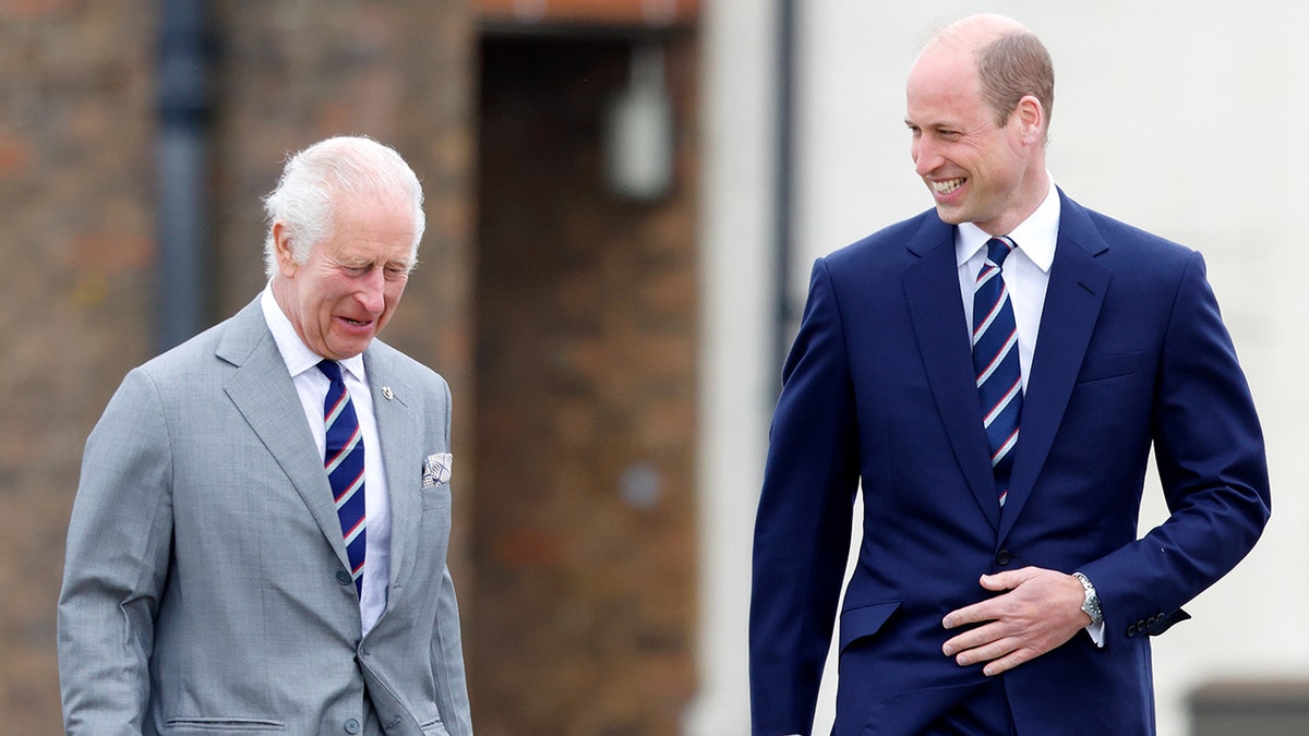 King Charles in a grey suit smiles and looks down as he walks with son Prince William in a navy suit