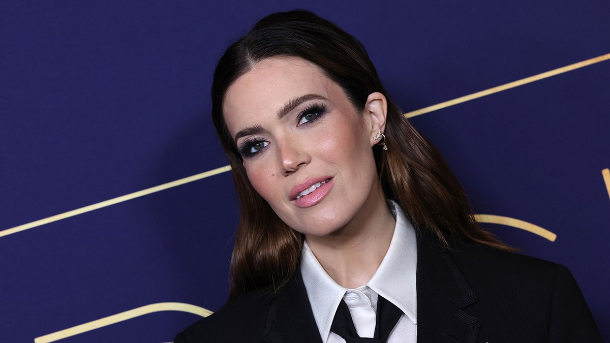 Mandy Moore con chaqueta negra, corbata y camisa blanca abotonada mira a la cámara