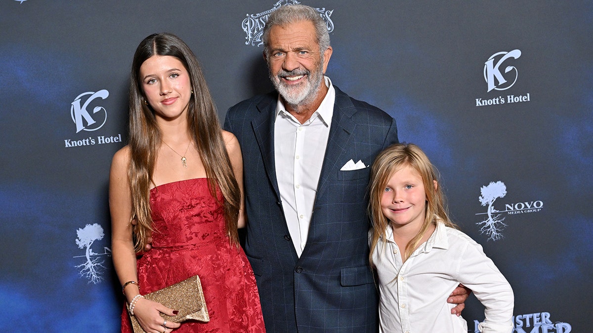 Mel Gibson, con un traje azul, posa en la alfombra con su hija Lucia, con un vestido rojo de tirantes, y su hijo Lars, con una camisa blanca y pantalones negros.