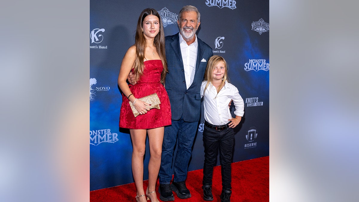 Mel Gibson, con un traje azul, sonríe en la alfombra con su hija Lucia, con un vestido rojo de tirantes, y su hijo Lars, con una camisa blanca y pantalones negros.