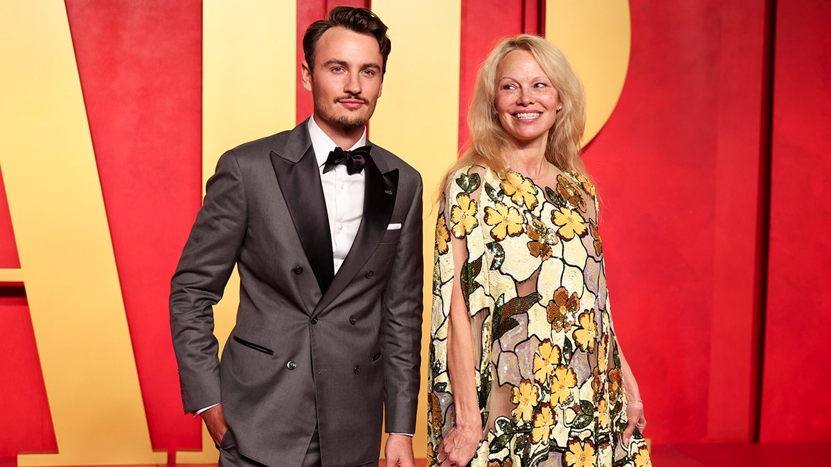 Brandon Thomas Lee in a brown/grey tuxedo with his mother Pamela Anderson in a yellow one-shoulder patterned dress at the Vanity Fair Oscar Party