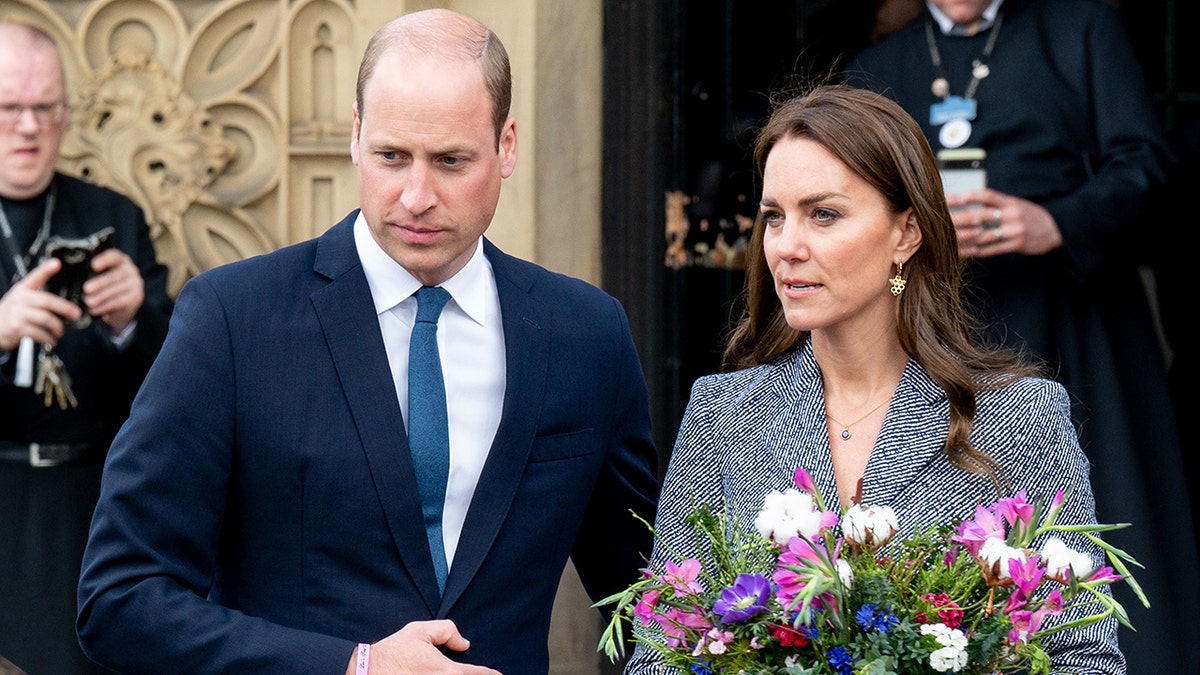 Prince William in a navy suit ushers wife Kate Middleton in a patterned suit holding a bouquet of flowers