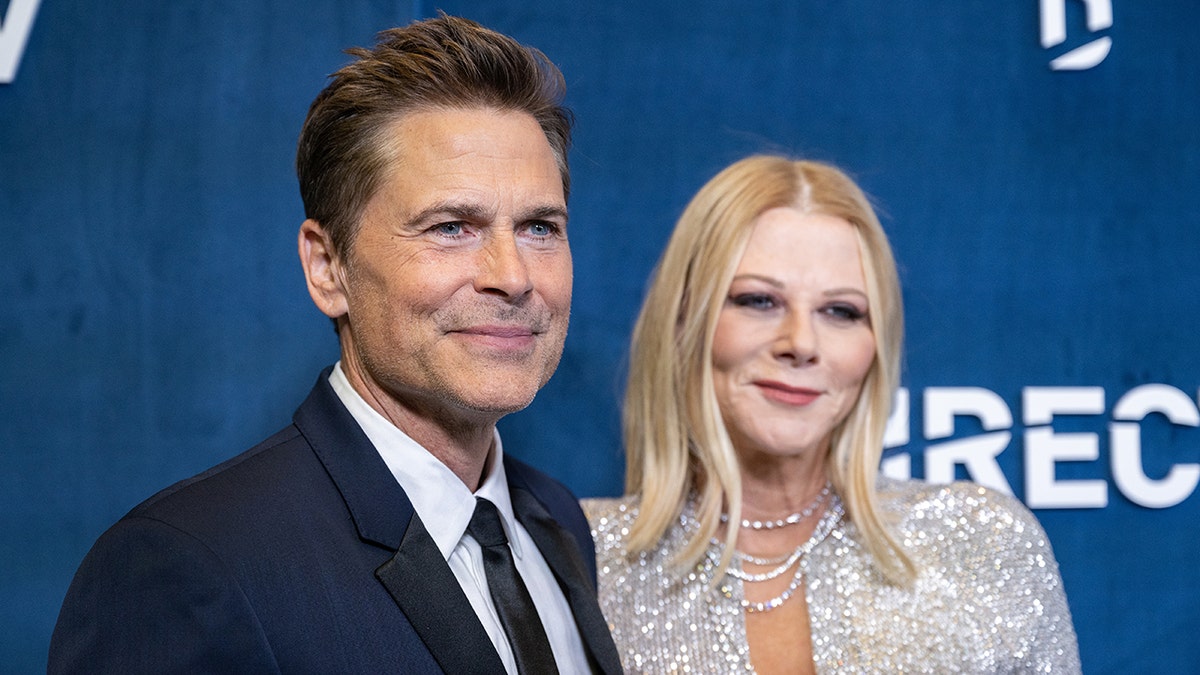 Rob Lowe con traje azul y corbata suave sonríe en la alfombra con un vestido plateado brillante