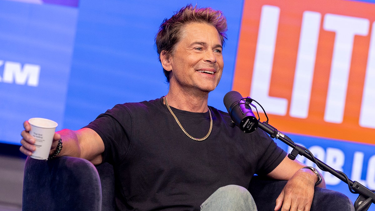 Rob Lowe sits in a chair, wearing a black shirt and gold chain, recording his podcast