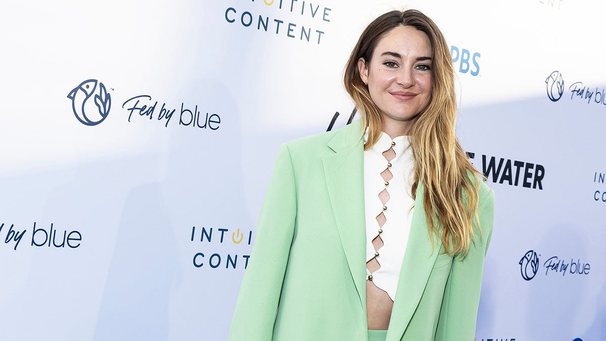 Shailene Woodley con una camisa blanca recortada y un blazer verde menta sonríe suavemente en la alfombra