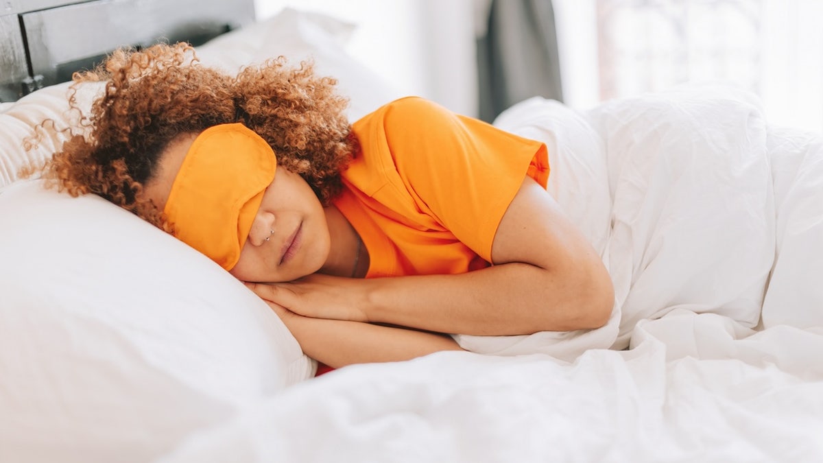 mujer con antifaz para dormir tumbada tranquilamente bajo las sábanas de la cama