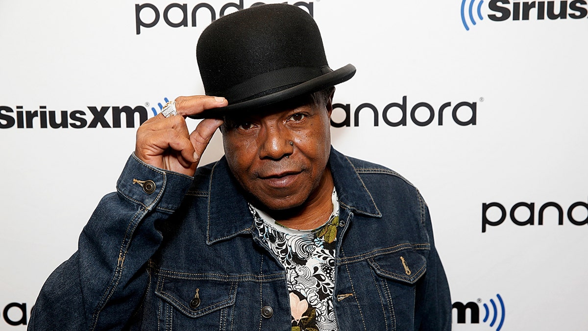 Tito Jackson mengenakan jaket jins melihat ke kamera saat dia menurunkan topi hitamnya