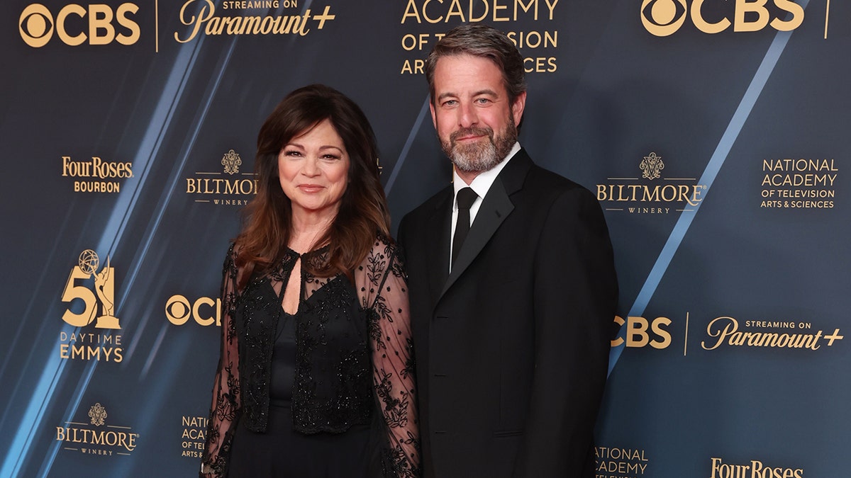 Valerie Bertinelli con un vestido negro de manga larga de encaje suave sonríe en la alfombra con su novio Mike Goodnough con un traje negro