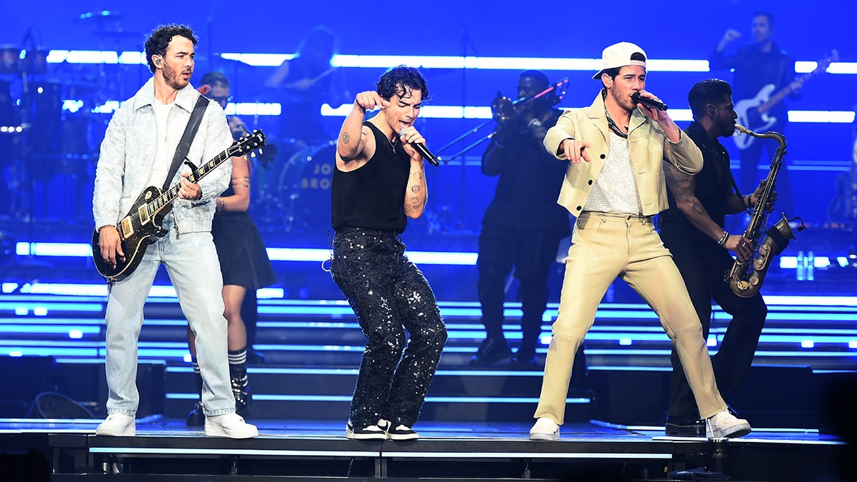 Kevin Jonas in a light denim jacket and pants, Joe Jonas in a black tank top and leather pants and Nick Jonas in a yellow suit perform on stage