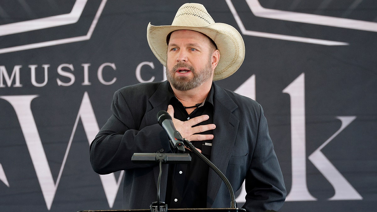 Garth Brooks puts his hands to his thorax  down  a podium wearing a achromatic  suit   and tan cowboy hat