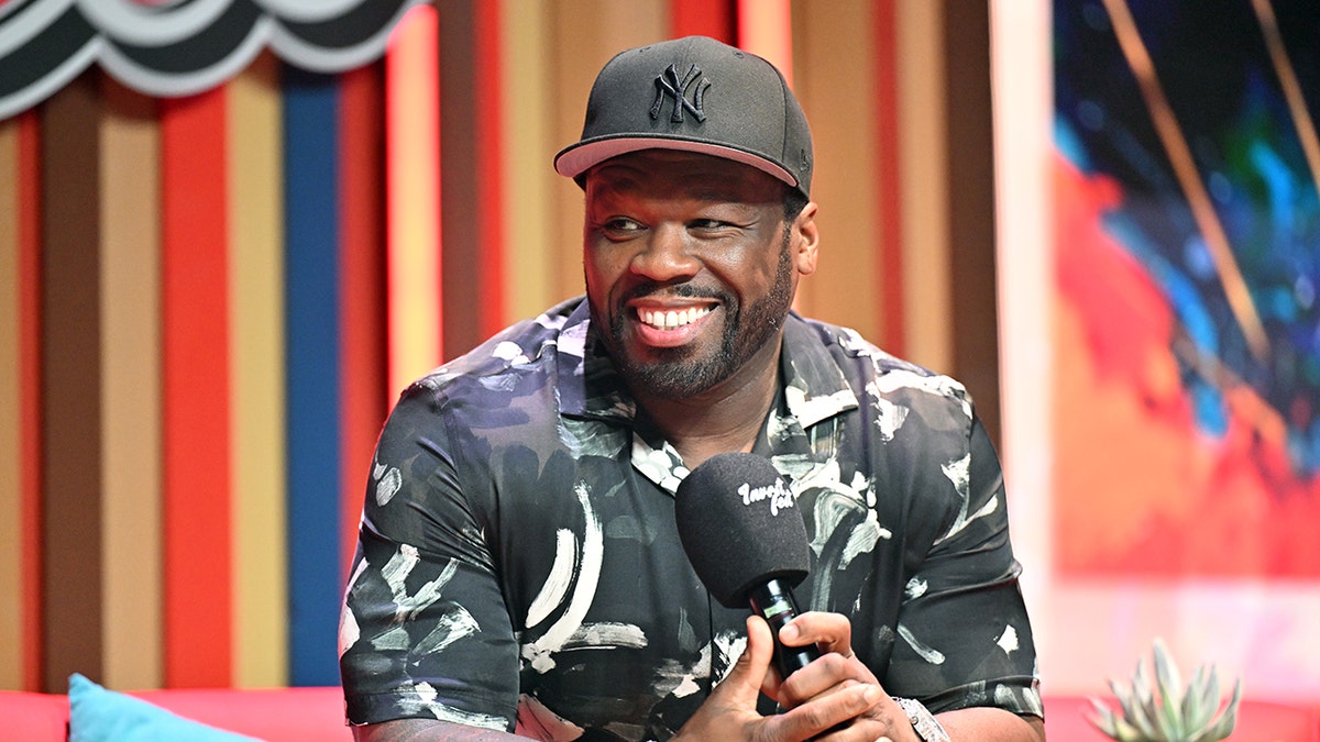 50 cent con una gorra de béisbol negra de los New York Yankees y una camiseta estampada sonríe sujetando un micrófono en el escenario