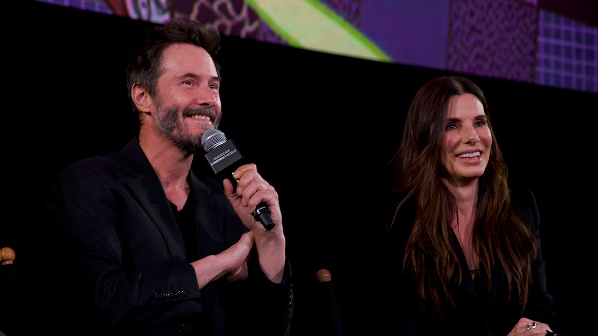 Keanu Reeves smiles connected  signifier    arsenic  helium  holds a microphone to his rima  sitting adjacent  to smiling Sandra Bullock, some  successful  black