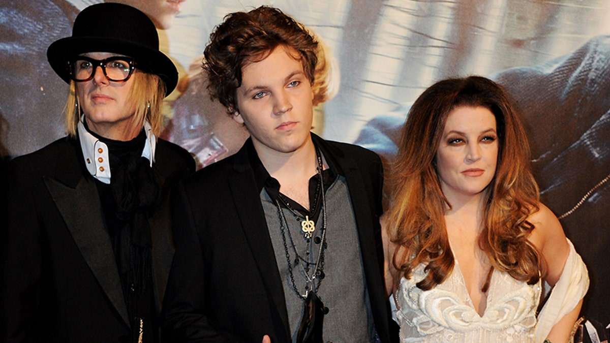 Michael Lockwood in a black jacket and hat lies on the carpet, stepson Benjamin Keough in a black jacket stands next to mother Lisa Marie Presley in a white dress